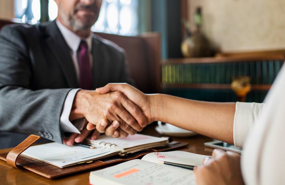 A paralegal shaking hands with their client over notes and documents in a lawyer's office.