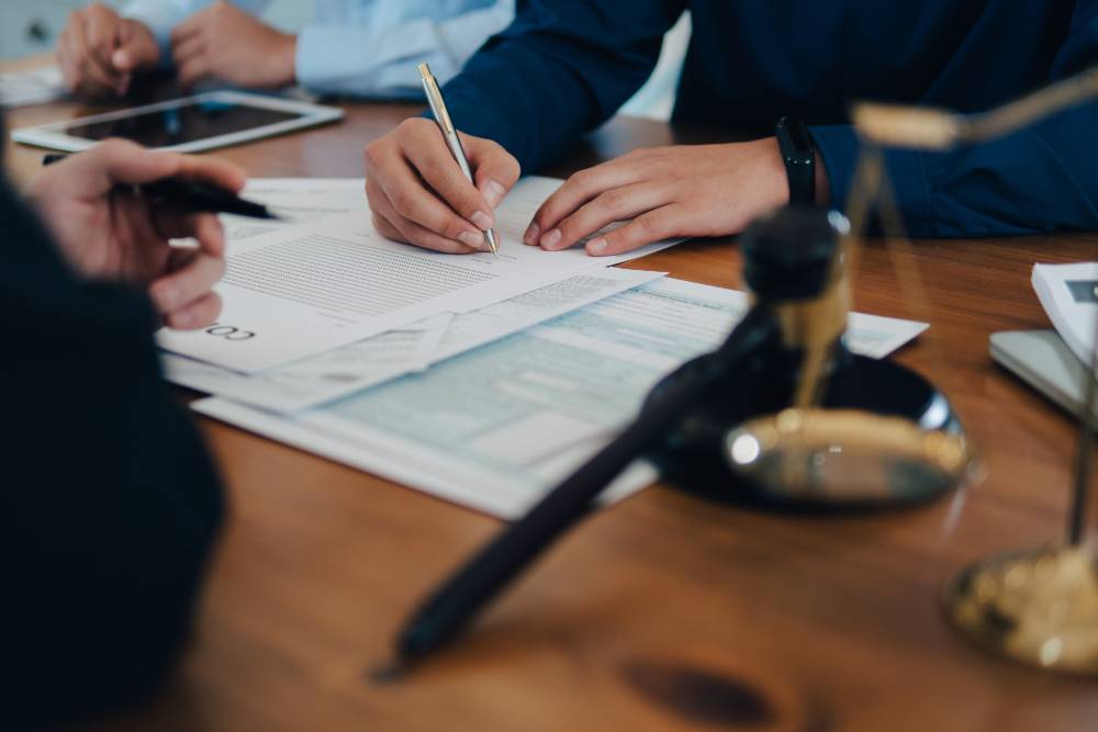 Three people at a desk reviewing law documents with pens as one of them is signing one of the documents.
